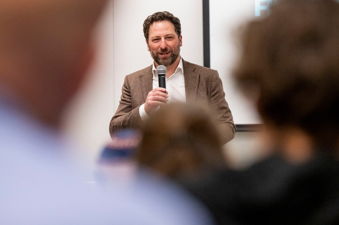 A man in a brown blazer and white shirt speaking into a microphone to an audience, with attendees visible in the foreground out of focus.
