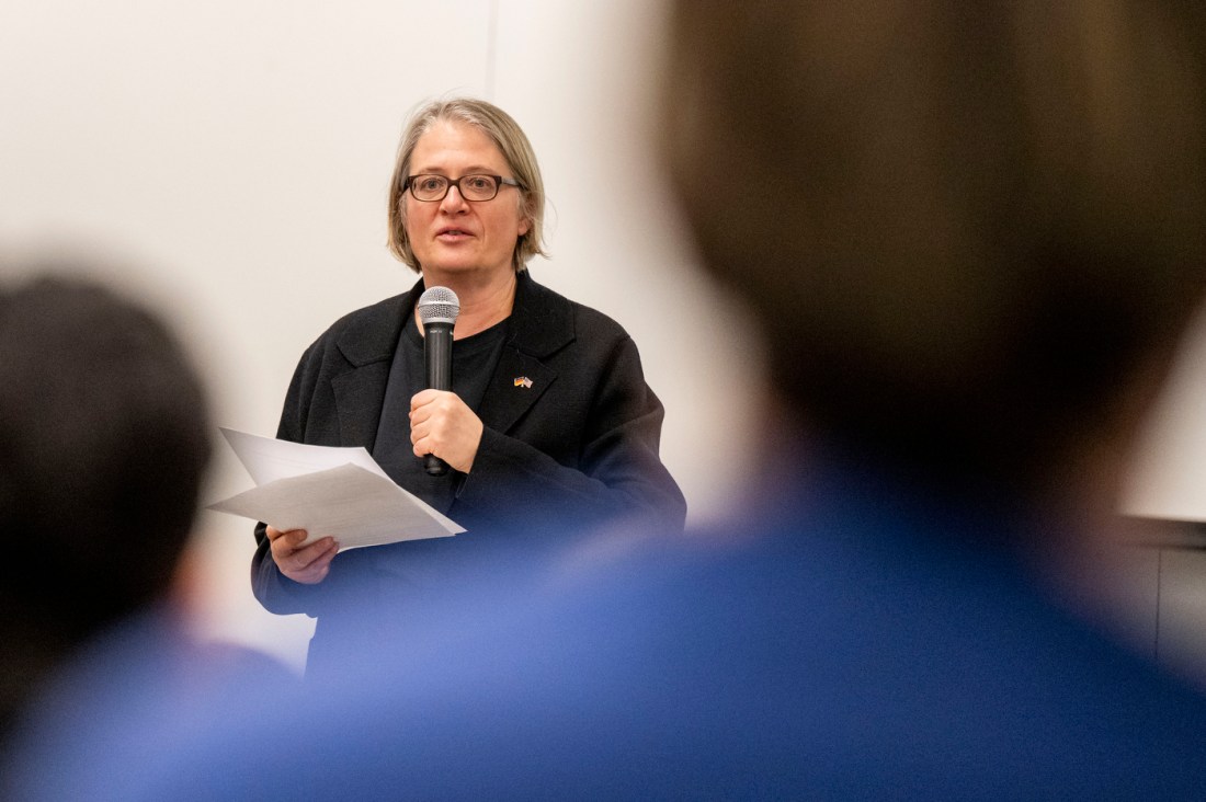 A woman with glasses wearing a black top speaking into a microphone while holding papers, with audience members visible in the foreground out of focus.
