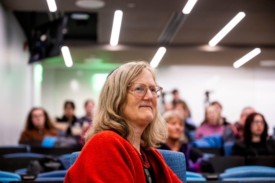 Rabbi Sara Paasche-Orlow in profile wearing a red cardigan, glasses, and a medal on a ribbon, seated in an auditorium with audience members visible in the background.