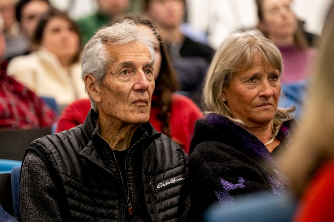 Two audience members, a man in a black jacket and a woman in a purple sweater, sitting attentively in a crowd with other attendees visible in the background.