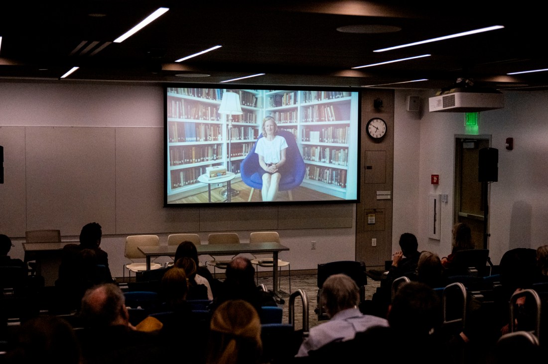 An audience seated in a dark auditorium watching a large projection screen displaying a video of Rabbi Sara Paasche-Orlow sitting in a room speaking, with floor-to-ceiling bookshelves in the background.