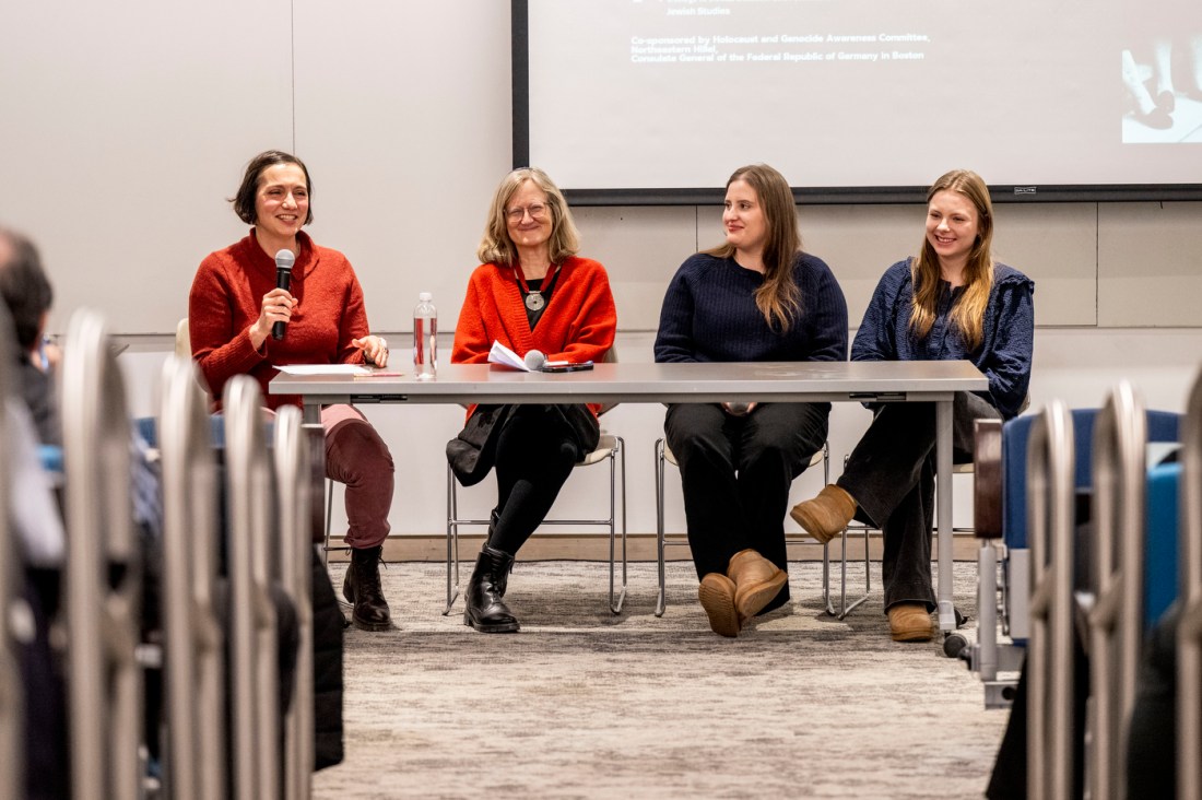 Four panelists seated at a table on stage—two women in red cardigans on the left, and two women in dark clothing on the right—with a projection screen behind them and audience members visible in the foreground.