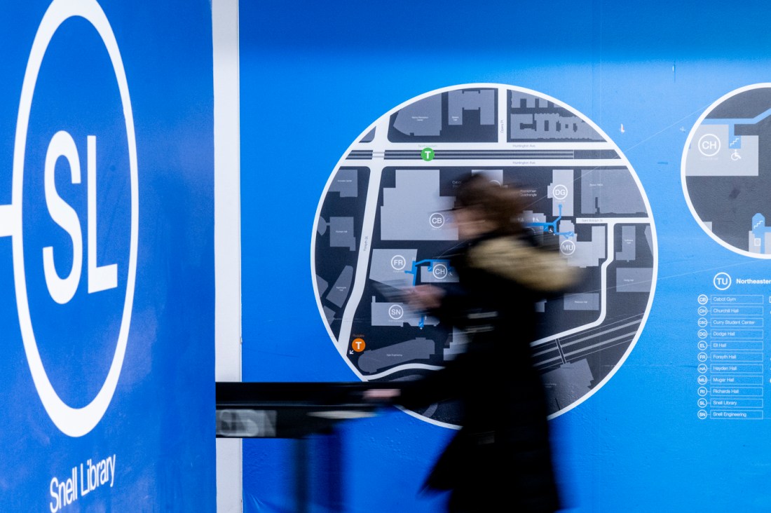 A blurred student walks past a bright blue wall featuring a logo that has 'SL' in a white circle. 