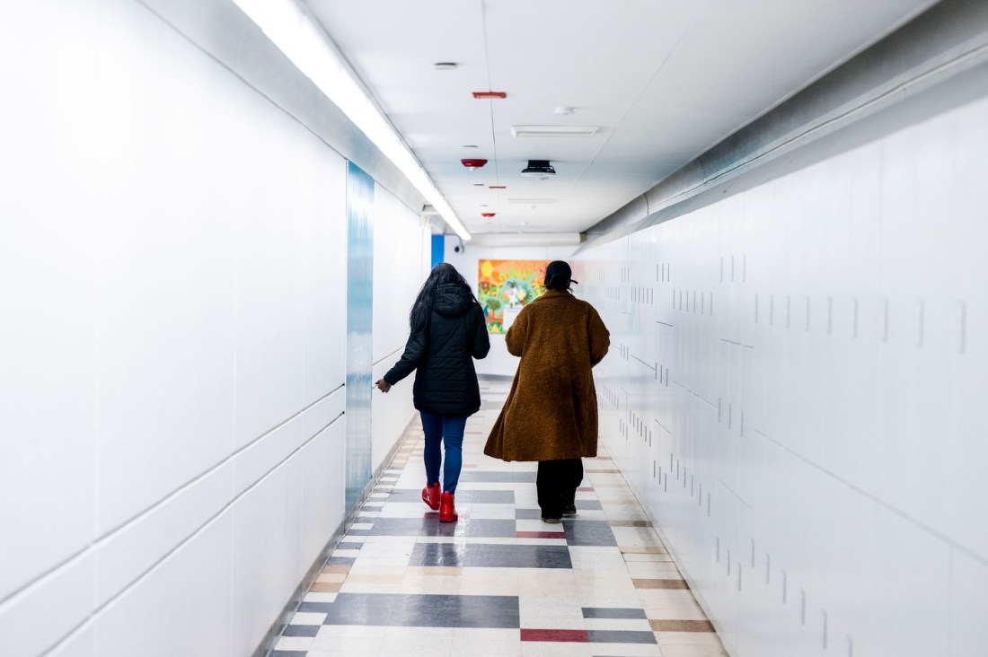 Two students in winter coats walk down a bright, minimalist hallway with white walls, white lockers, and polished floors. 