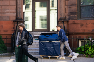 Two students walking on a sidewalk, one pushing a cart full of items they're moving into their off-campus housing.