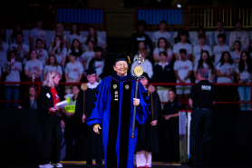 A woman dressed in a purple-and-black academic robe with golden regalia, a black cap with a golden tassel and carrying a wooden mace in her left hand leads the cadre of marshals during an academic ceremony.