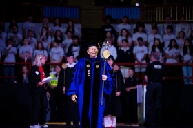 A woman dressed in a purple-and-black academic robe with golden regalia, a black cap with a golden tassel and carrying a wooden mace in her left hand leads the cadre of marshals during an academic ceremony.