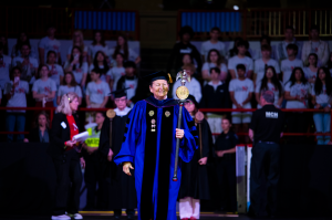 A woman dressed in a purple-and-black academic robe with golden regalia, a black cap with a golden tassel and carrying a wooden mace in her left hand leads the cadre of marshals during an academic ceremony.