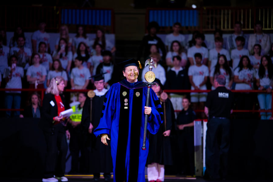 A woman dressed in a purple-and-black academic robe with golden regalia, a black cap with a golden tassel and carrying a wooden mace in her left hand leads the cadre of marshals during an academic ceremony.