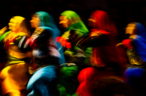 Blurry silhouettes of five women dance in colorful outfits — yellow, light blue, green, red and sapphire blue.