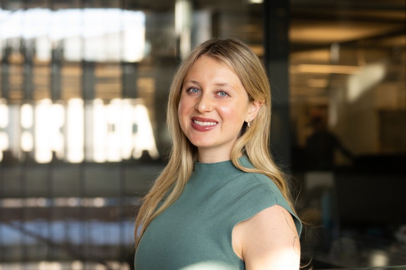 Kristen Kilgallen with blonde hair in a short-sleeved green top smiling at the camera in a modern interior space with bright lights visible in the background.