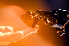 A robot hand touching one finger to the finger of a human hand. The photo is overlaid with orange lighting.