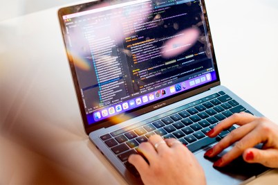A person's hands, with red painted nails, at work on a laptop displaying data or strings of code.