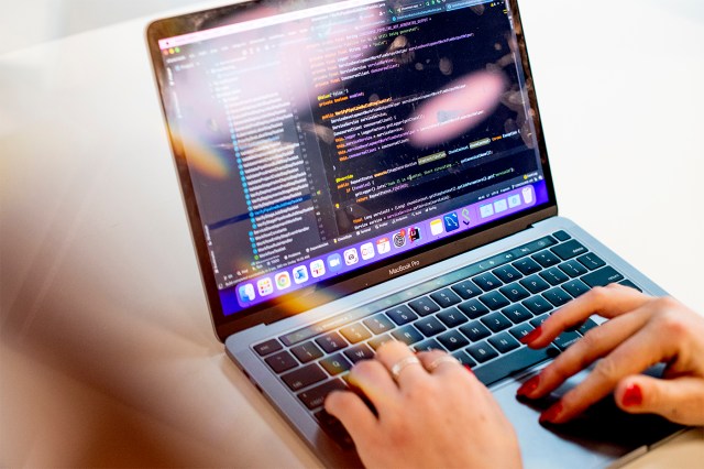 A person's hands, with red painted nails, at work on a laptop displaying data or strings of code.