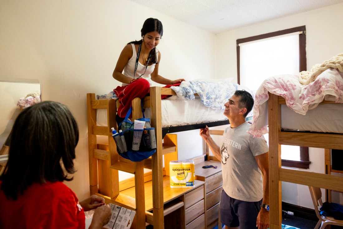 Grace Benavidez sitting on top of a twin bunk bed with her parents standing in her dorm next to the bed.