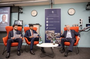 Three suited men sat on orange chairs during a presentation