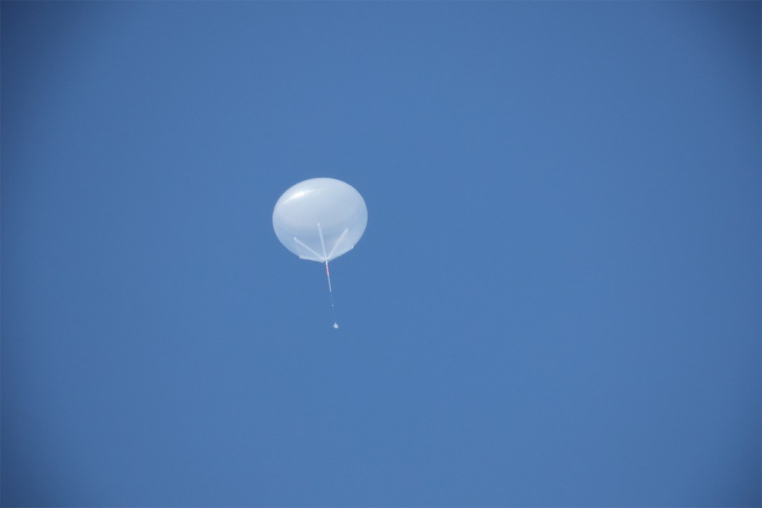 A large weather balloon floats very high in the sky. Below it, one can just make out the payload hanging from its tether.