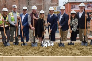 A group of people wearing hard hats pose for a photograph as they break ground on a section of ground with shovels.