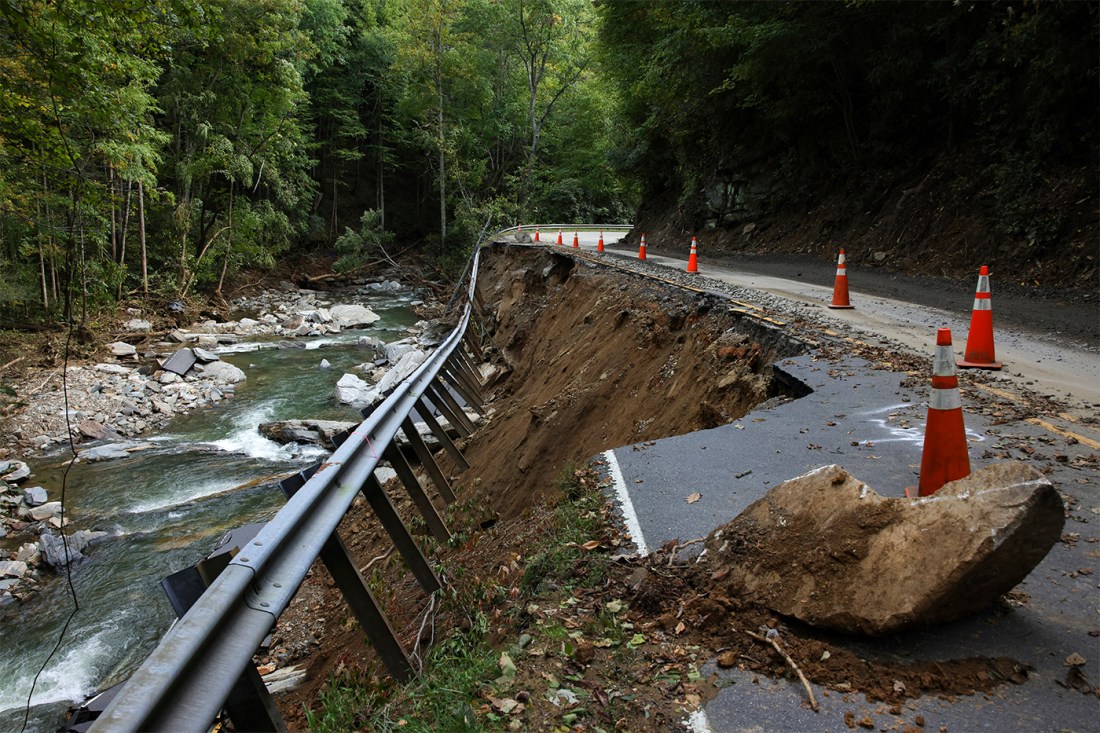 A river rushes by a damaged road with cones on it in Asheville after Hurricane Helene.