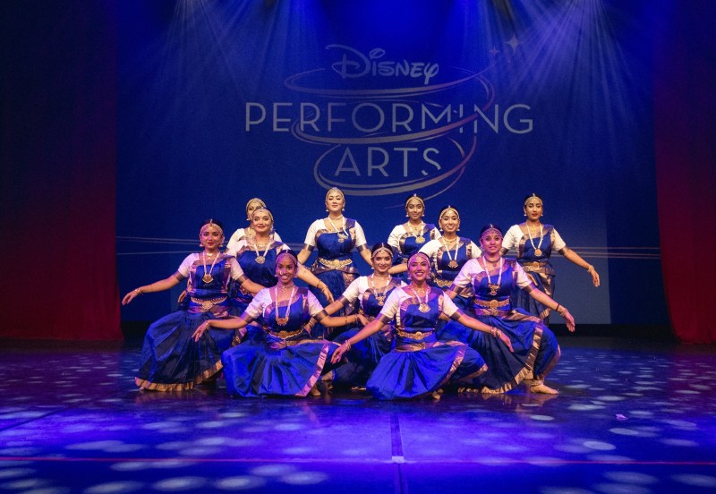 A group of dancers in traditional blue and gold costumes pose in formation on a purple-lit stage, with 'Disney Performing Arts' backdrop visible behind them.