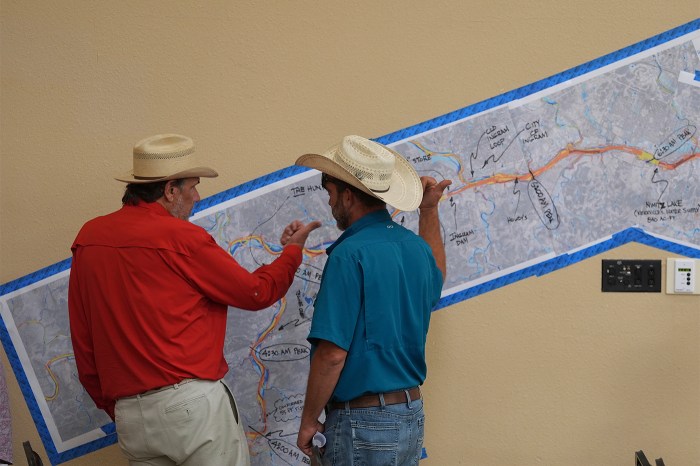 Two men wearing hats, one in a blue shirt and one in red, look at a map taped to a tan wall.