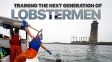 On an overcast day on the ocean, one person steers a small fishing boat while another pulls up a lobster trap. Text on the image says 'Training the next generation of lobstermen'. There is a lighthouse in the middle distance.