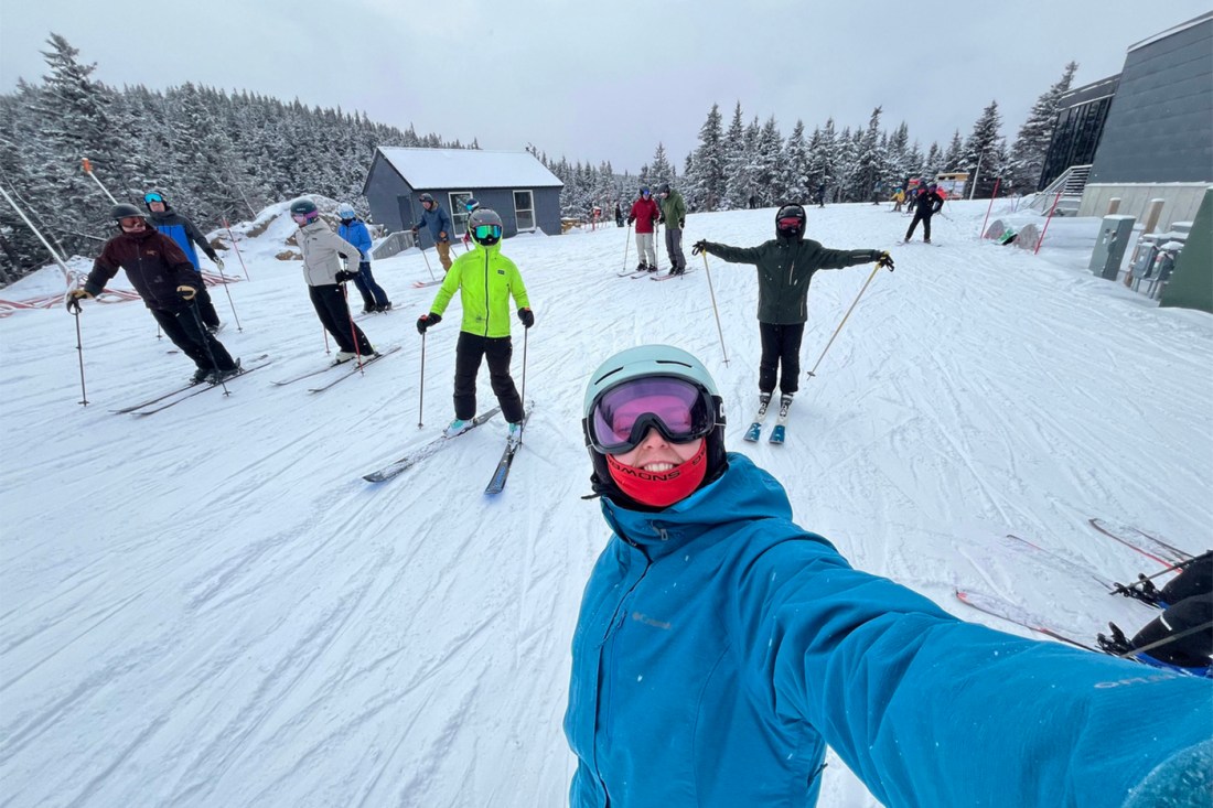 Members of the club in a ski area posing for a photo in their skiing gear and skies, while a person in the foreground takes a selfie with them.