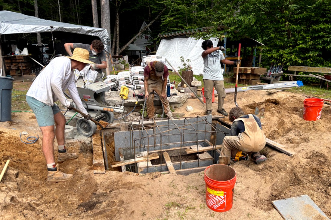 A group of NUHOC club members works on building a foundation pier for the lodge in the sand, surrounded by forest, in the summer, with tents in the background.
