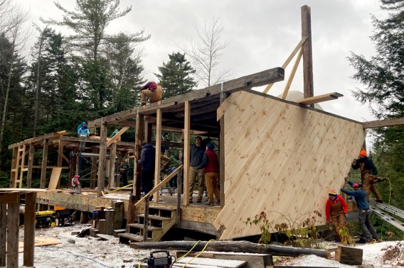 A photo of the wooden frame of the new lodge with partial sheating among fir trees and snow with people working on it.