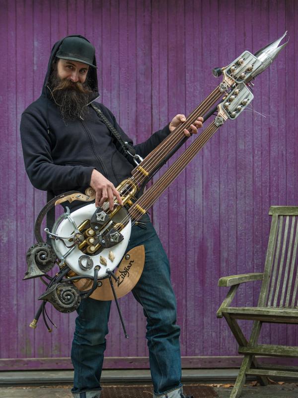 Michael Ulman poses in a black hoodie while holding the large, double-necked flame-throwing guitar he made for "Mad Max: Fury Road."