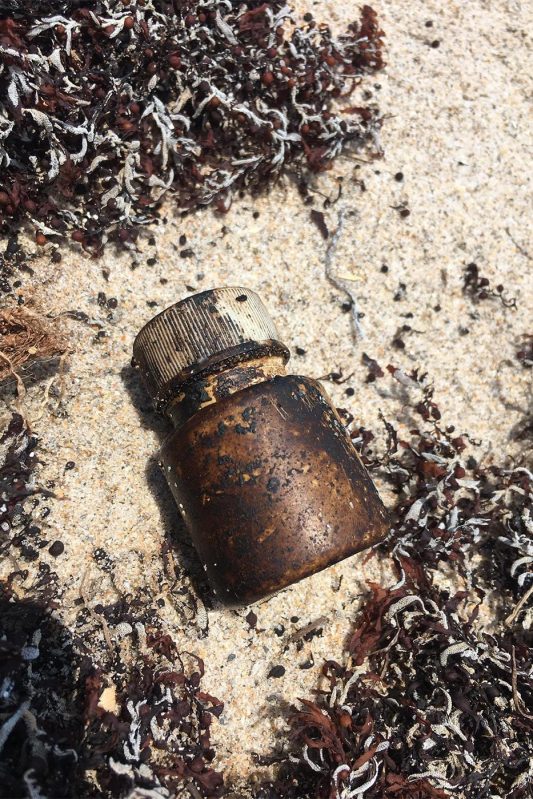 A plastic container with a screw top lid, contaminated with oil, lies in sand.