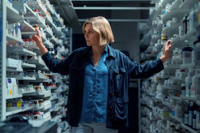 A woman dressed in blue denim stands between shelves containing pharmaceuticals and other bottles.