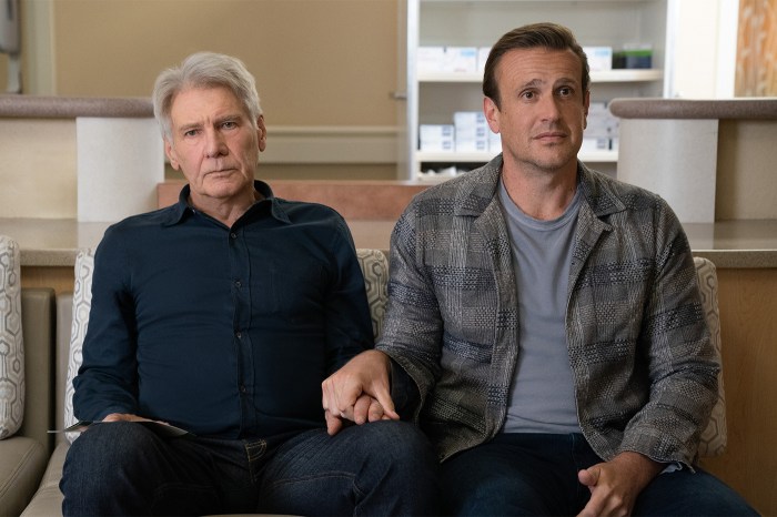 Two men sit side by side on a beige couch in a bright, modern interior with white cabinetry visible in the background, one wearing a dark button-up shirt and the other in a gray patterned jacket over a gray shirt.