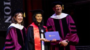 A smiling graduate flanked by faculty in ceremonial robes. They hold a blue diploma.