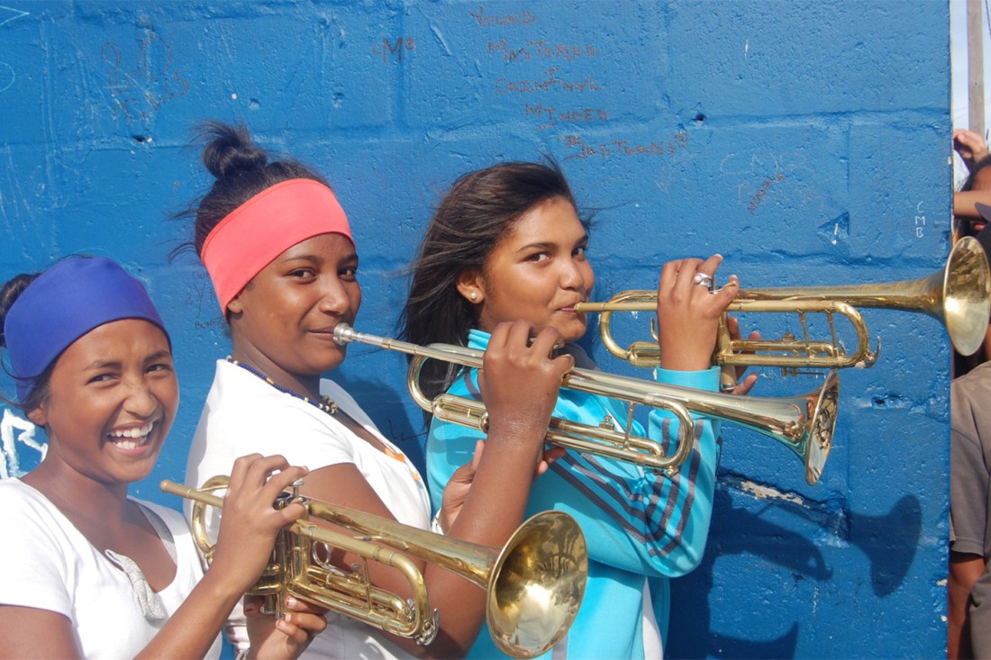 Three young women play trumpets, standing in front of a blue wall and looking into camera. One of them is laughing.