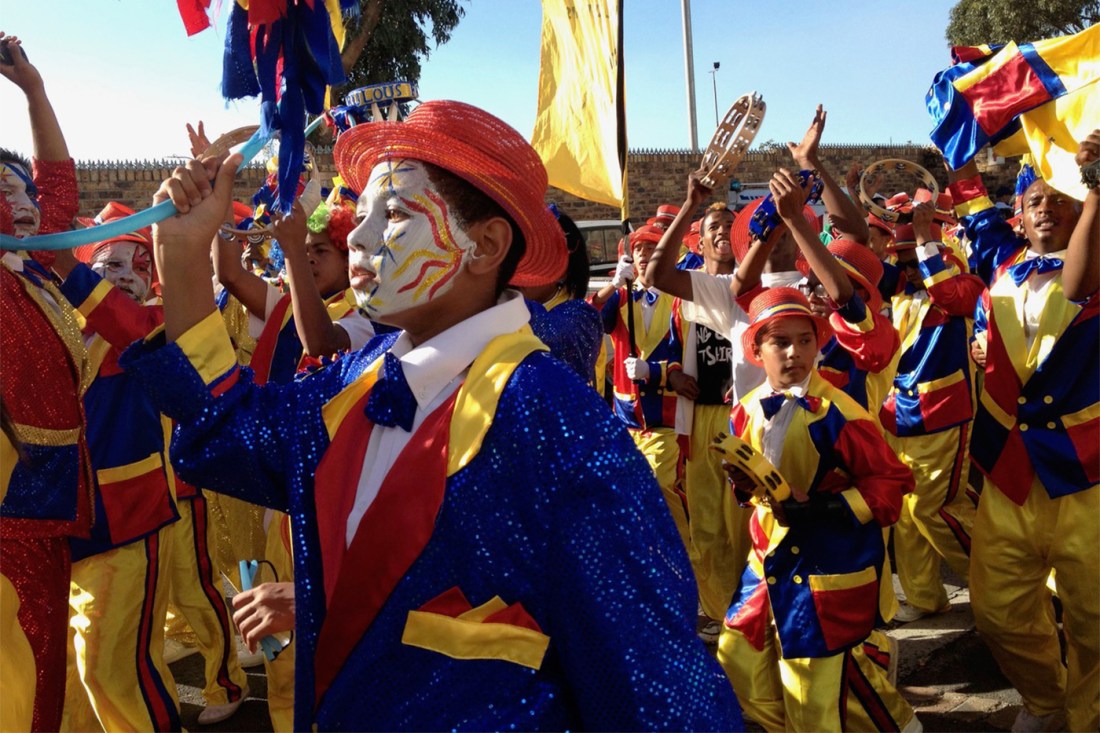 In the foreground, a Kaapse Klopse participant wears a blue suit, red hat and white face paint. Many other people surround him, wearing many colors, but predominantly yellow.