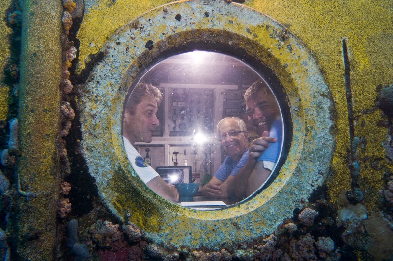 Researchers inside an underwater habitat visible through a circular porthole encrusted with yellow and green marine growth.
