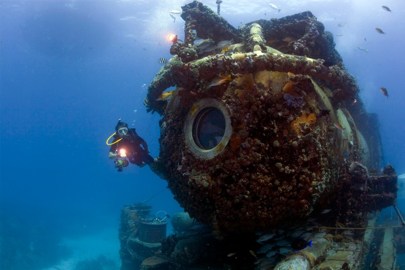 A diver swimming alongside a underwater research habitat covered in marine and coral growth, with blue ocean water and fish swimming in the background.