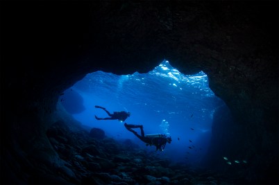 The silhouettes of two scuba divers in a patch of blue water surrounded by a dark cave underwater.