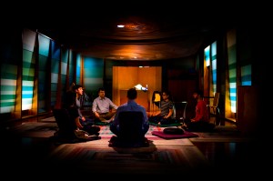 A group of people sit with their legs crossed in a dimly lit meditation center.