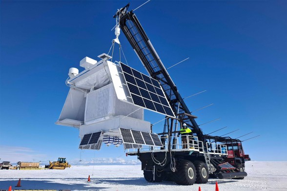 A crane holds a large white box with solar panels on the side. The box hangs above snow or ice-covered ground, and is framed by blue sky.