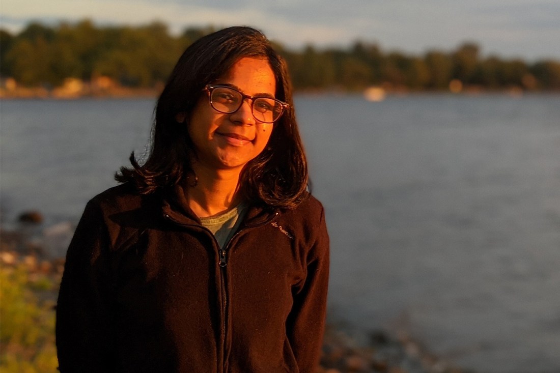 A woman with long hair, lit by the sunset, stands in front of a lake.