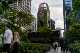 A modern elevated building with vertical gardens stands among downtown high-rise buildings, with pedestrians walking along a tree-lined pathway below.