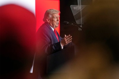 U.S. President Donald Trump speaking while gesturing with a hand in front of a red backdrop.