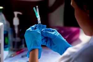 A health professional with gloved hands holding up a syringe and looking at it.