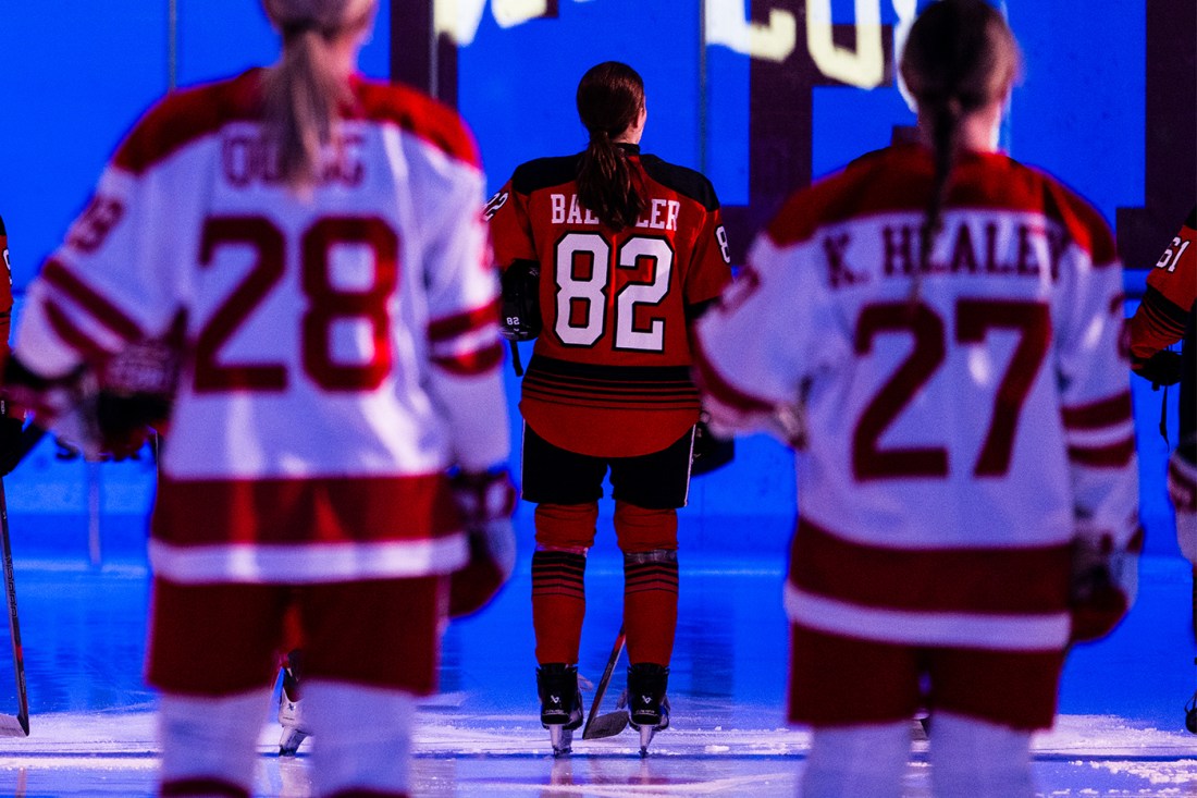 Alessia Baechler standing on the ice wearing a red jersey that has her last name and the number '82' on the back. Blurred in the foreground are team members from the opposing team wearing white jerseys. The arena they are in is lit with blue lighting.