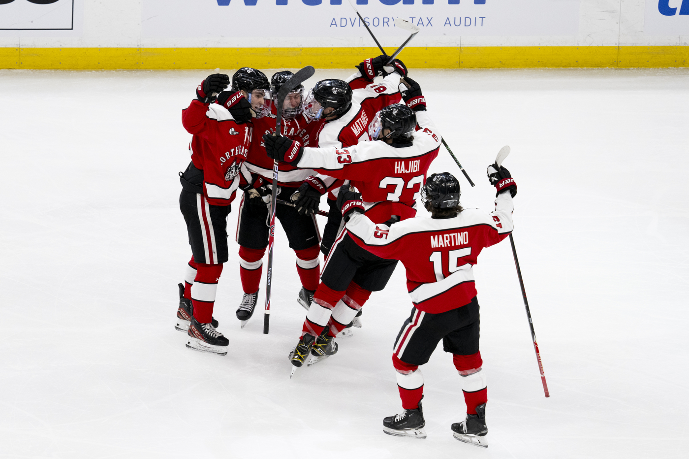 Northeastern men’s hockey players in red jerseys cluster together in celebration on the ice after a goal during the Beanpot semifinal.
