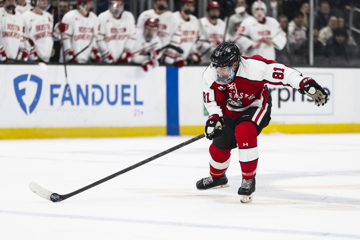 A Northeastern player wearing a red No. 81 jersey skates forward with the puck as Boston University players watch from the bench in the background.