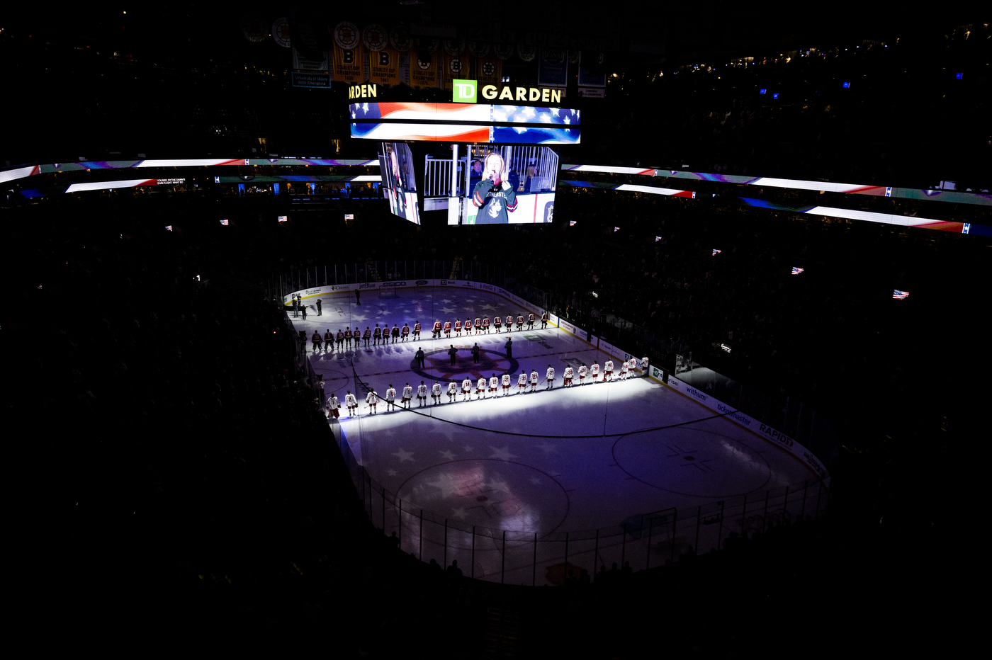 The Boston University Terriers and the Northeastern Huskies stand opposite each other on the ice with the stadium dark.