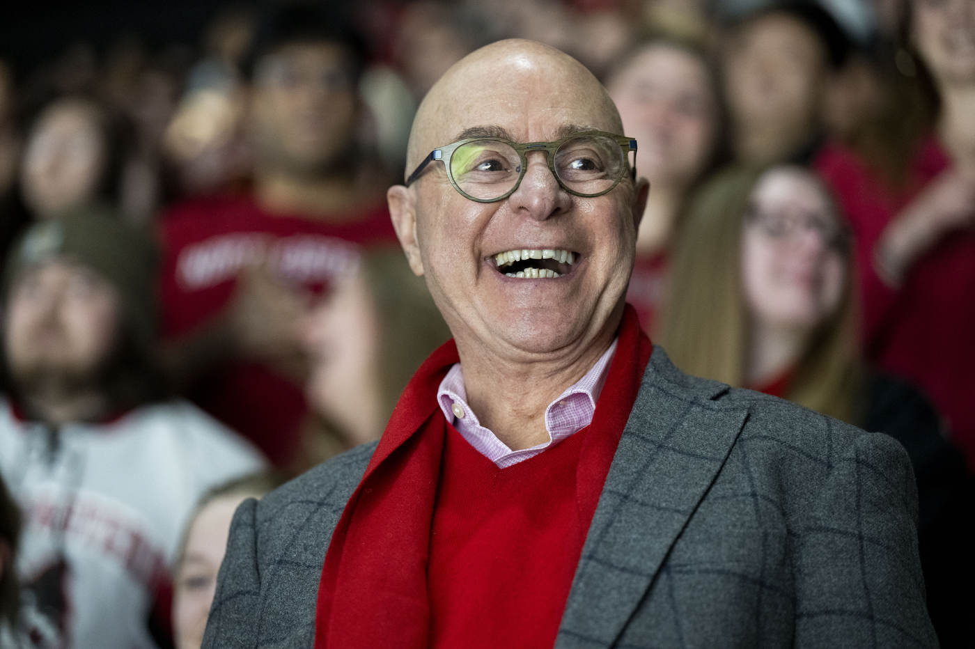 Northeastern University President Joseph Aoun wearing glasses, a gray blazer, and a red scarf looks on from the stands, surrounded by cheering fans in red and black.